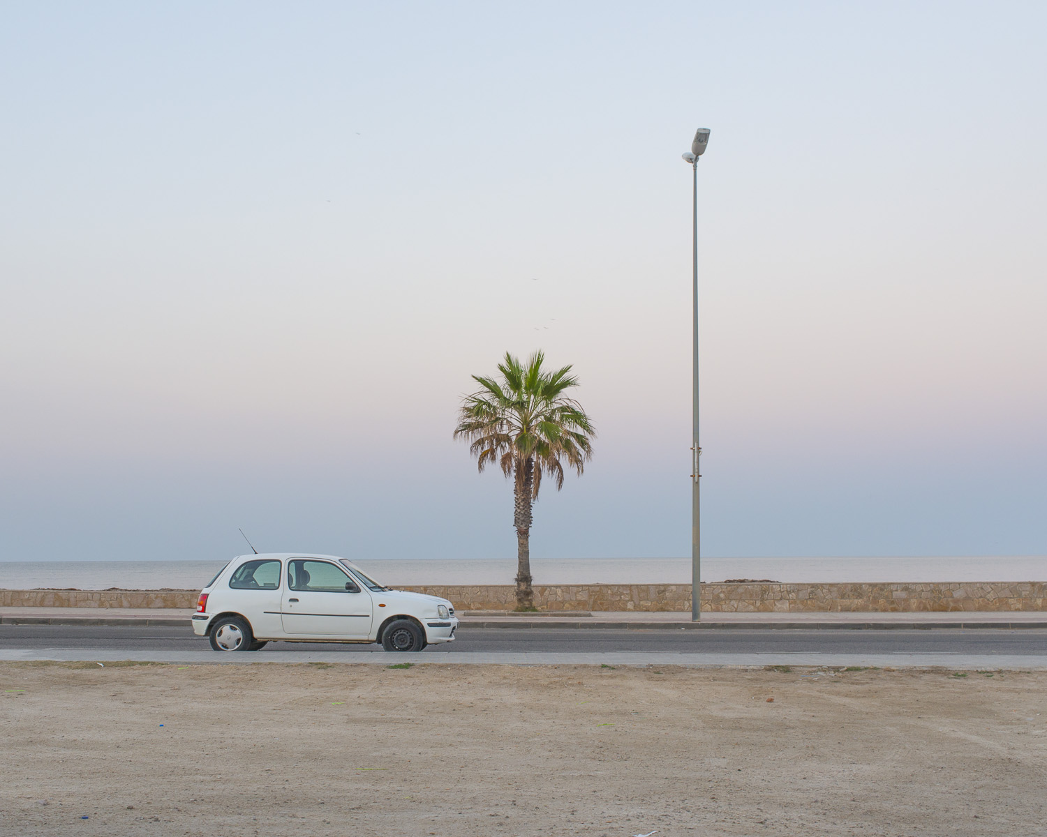 A white car stands on a straight, smooth road, surrounded by soft sand and a graceful palm tree and lamppost. The colors are light and tranquil, and the emptiness of the surroundings creates space for reflection and freedom. It is a beautiful and serene composition that invites us to escape the busyness of everyday life and enjoy the moment.